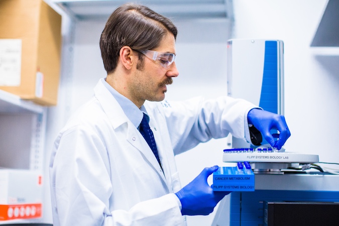 Profile view of man in safety goggles, lab coat, and purple gloves handling tubes in laboratory.