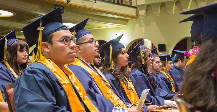 Graduates seated during commencement listen to a speaker on stage.